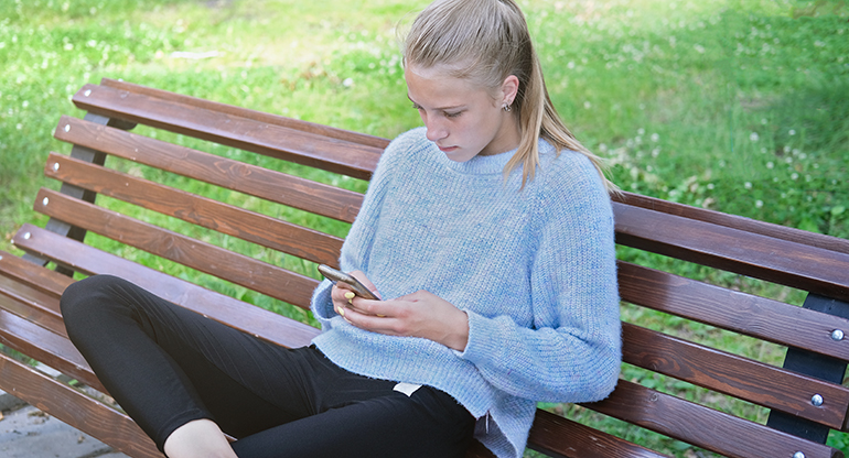 En tjej i tonåren sitter på en bänk och tittar på sin telefon. Foto.
